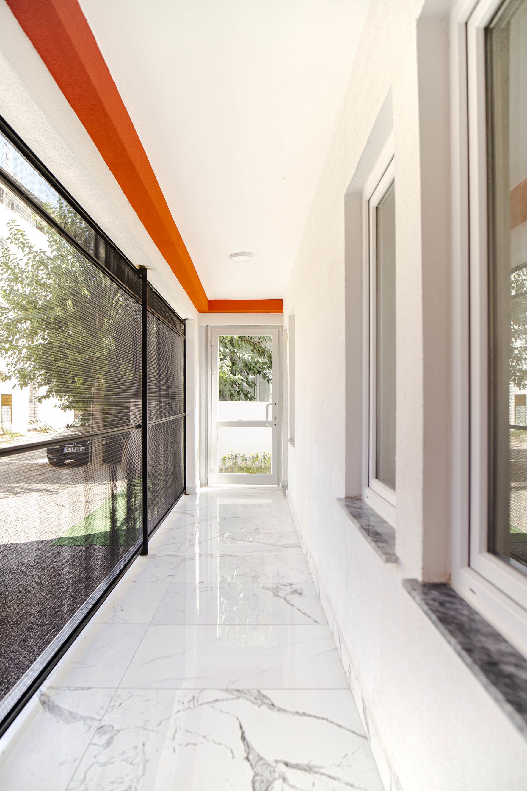 A corridor with a glass door leading to a balcony, a black mesh screen on the left, and a white wall with a marble floor.