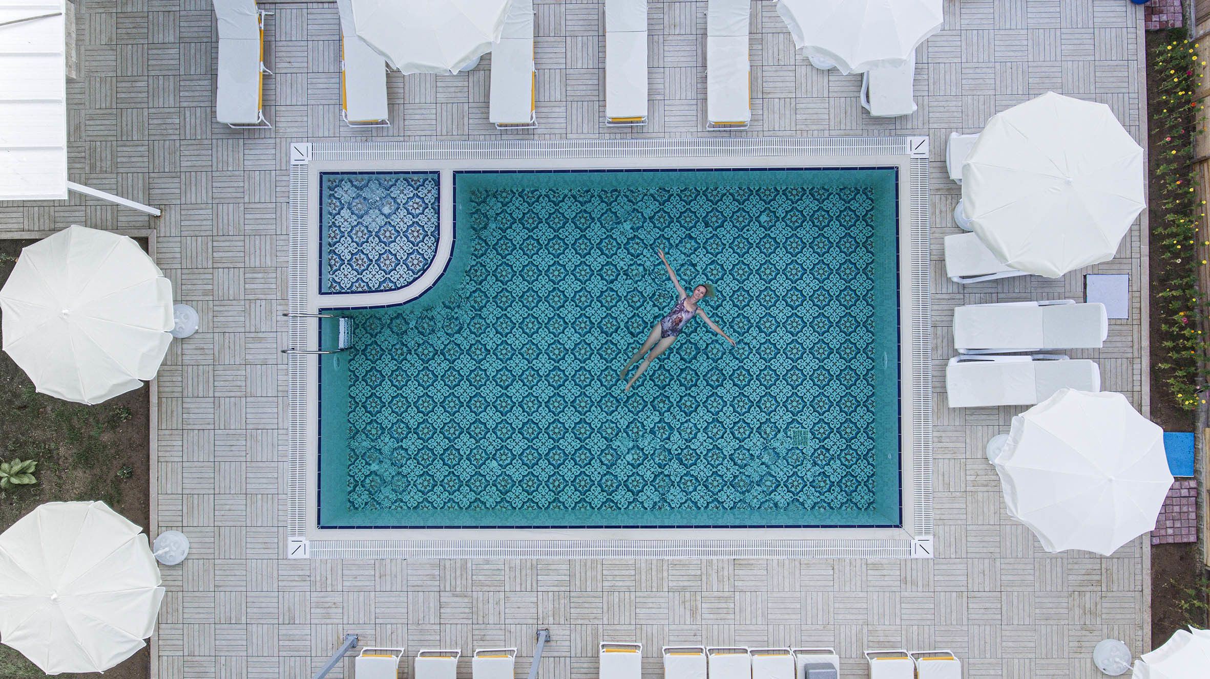 An aerial view of a rectangular swimming pool with a patterned floor, surrounded by white lounge chairs and umbrellas.