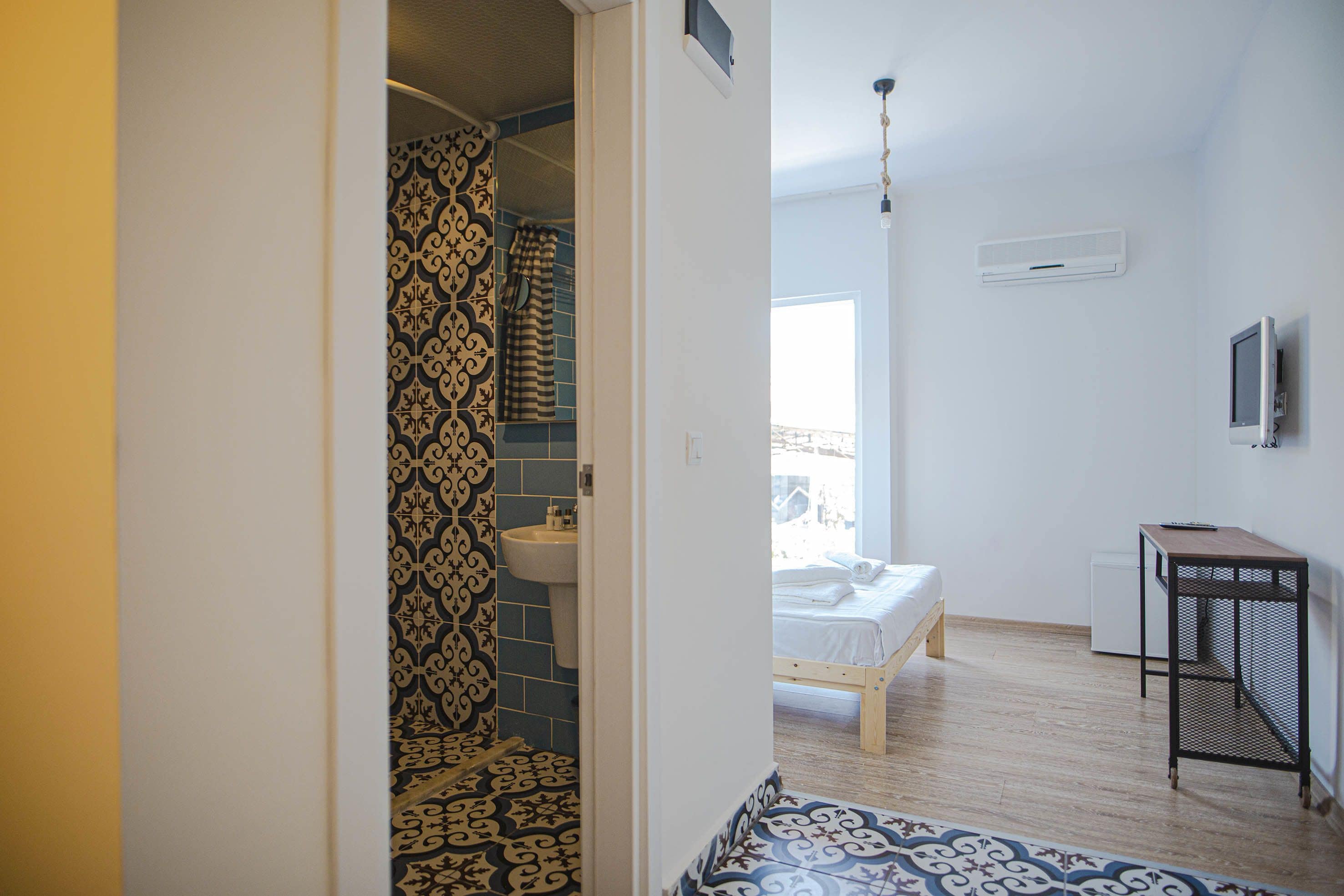 A bedroom with a bed, a bedside table, a TV, and a bathroom with a sink and patterned tiles.