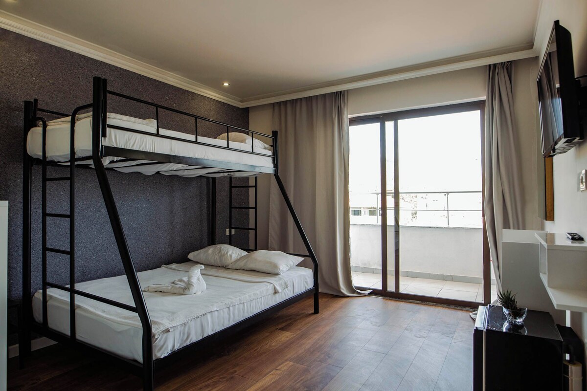 Une chambre avec un lit superposé, un balcon, une télévision et un bureau agrémenté d’une plante.