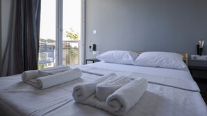Een net opgemaakt bed met witte lakens en opgerolde handdoeken, een raam met gordijnen en uitzicht op de omgeving.