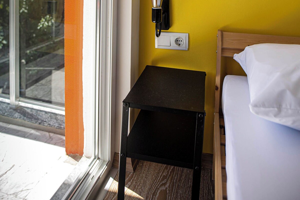 Une chambre d’hôtel avec un lit, une petite table, une prise électrique et un interrupteur.