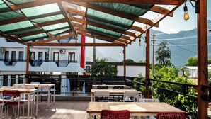Een terras buiten met houten tafels en rode stoelen, een pergola met een groen dak, en een uitzicht op gebouwen en bergen op de achtergrond.