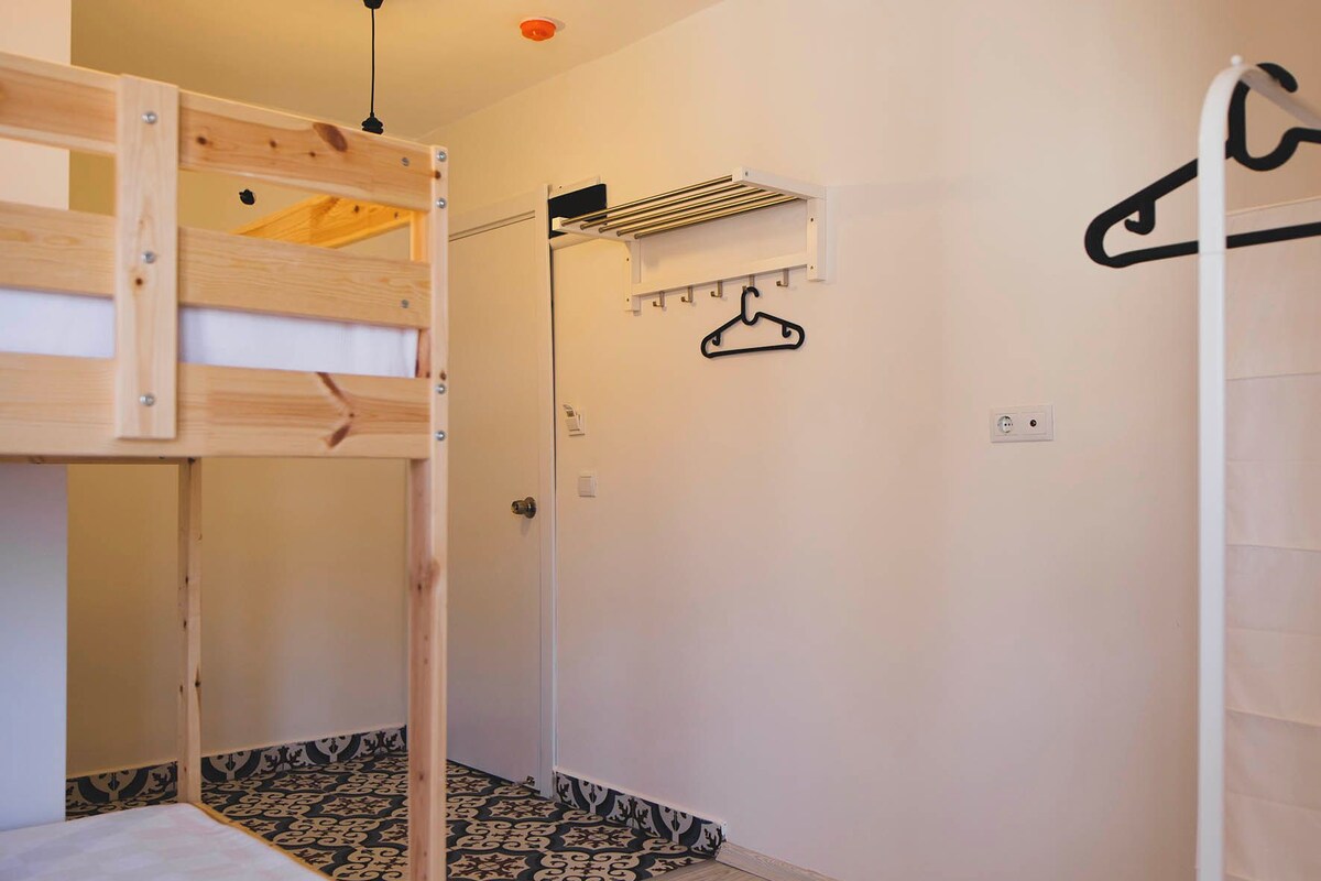 Un lit superposé avec une structure en bois et un matelas, un mur blanc avec un portant à vêtements suspendu et un tapis à motifs sur le sol.