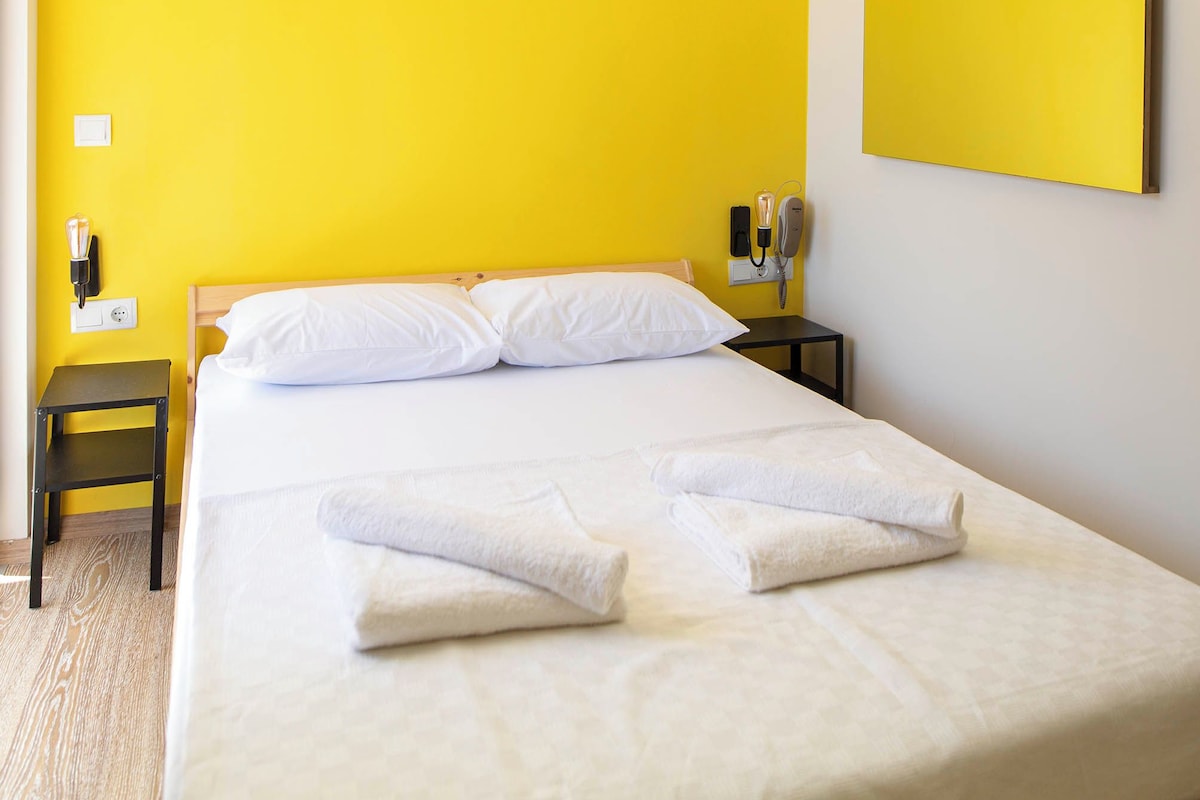 A hotel room with a large bed, white bedding, a yellow accent wall, a bedside table with a lamp, and a wall-mounted telephone.