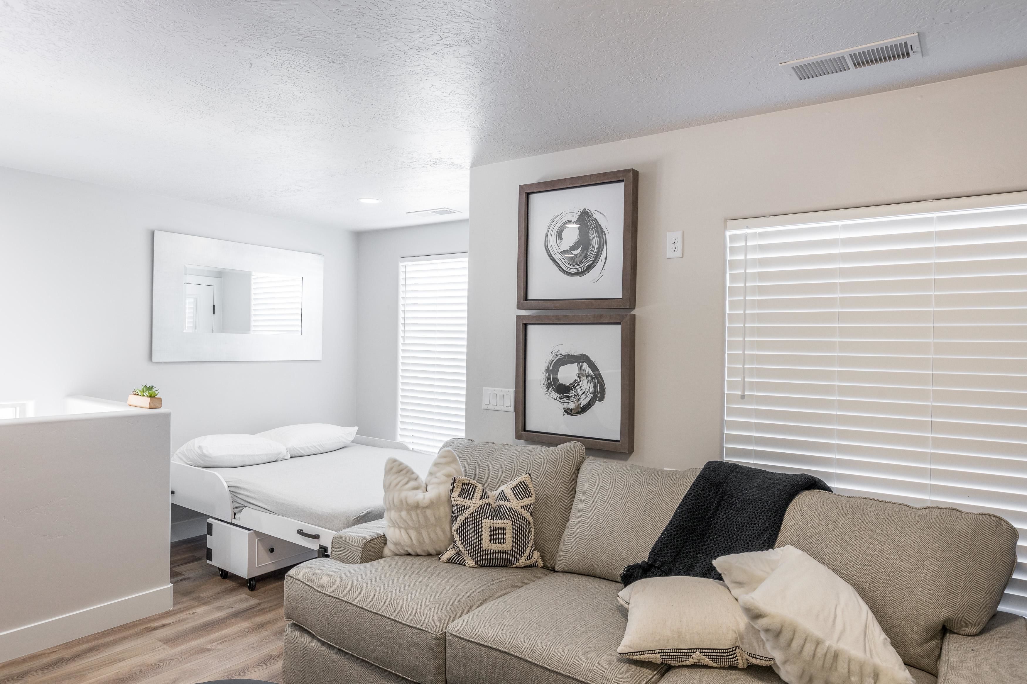 A modern, well-lit room with a beige sofa, a bed, and a small desk. There are two framed artworks on the wall and a window with blinds.