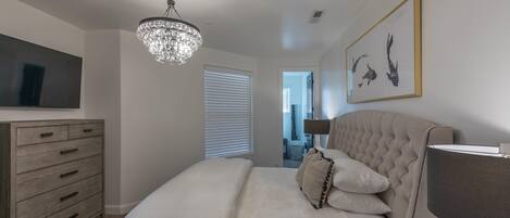 A bedroom with a bed, dresser, chandelier, artwork, and a television.