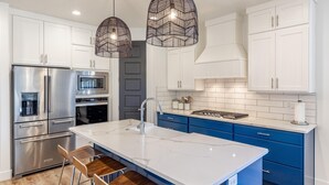 A modern kitchen with white cabinets, blue drawers, a stainless steel refrigerator, an oven, and a large island with a sink. - Fable House (St. George)