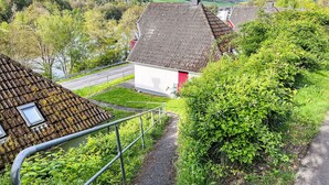 Una casa rural con techo inclinado, un pequeño jardín y un camino que lleva a una carretera.