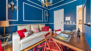 A room with a blue accent wall, white sofa with red and patterned cushions, a wooden dining table set for two, and a chandelier. - Modern stylish studio in downtown Lisbon center - new to Vrbo (Lisboa)