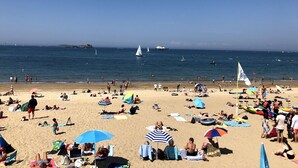 Ein belebter Strand mit badenden und sonnenden Menschen, Booten im Wasser und einem klaren Himmel.