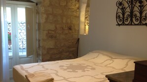 A bedroom with a stone wall, a bed with a patterned bedspread, a wooden dresser, and a decorative metal wall piece. - Casa San Pietro Modica (Modica)