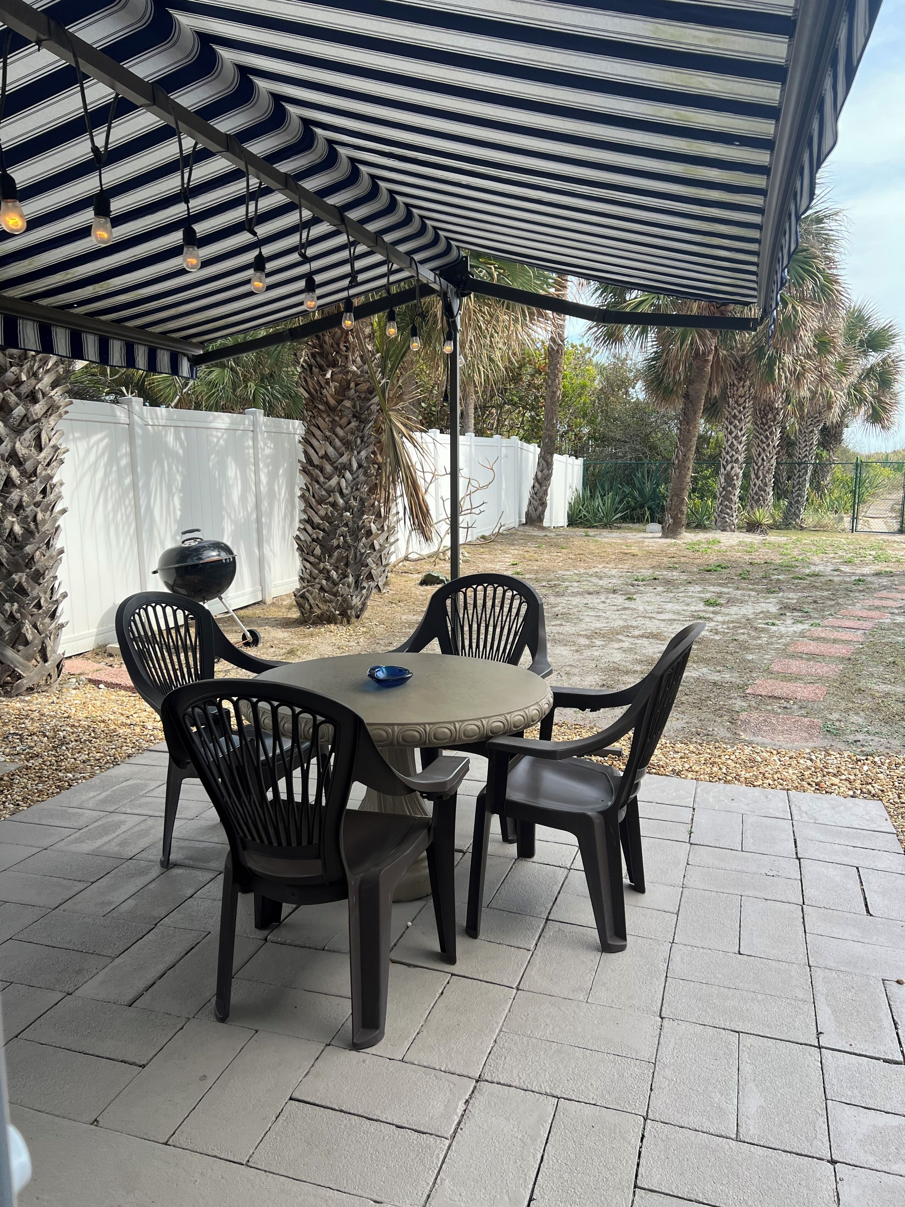 Une terrasse couverte avec une table et des chaises, un barbecue et, au fond, une clôture blanche.