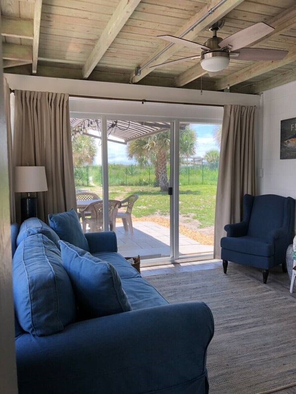 A living room with a blue sofa, a chair, and a table. There is a sliding glass door leading to an outdoor patio with a palm tree and a fence.