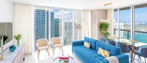 A modern living room with a blue sofa, a coffee table, and a dining area with a view of the cityscape.
