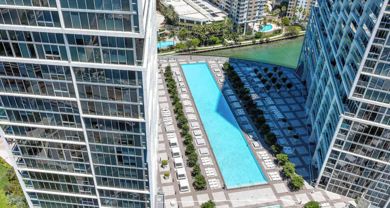 Un edificio alto con una piscina larga y estrecha rodeada de sillones de descanso y plantas en macetas.