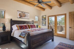 Cozy bedroom with a dark bed frame, colorful pillows, and a ceiling fan. A large window offers a view, and framed artworks adorn the walls.