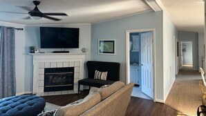 Ein Wohnzimmer mit Kamin, einem schwarzen Sofa, einem blauen Hocker und einem Flachbildfernseher, der über dem Kamin angebracht ist.