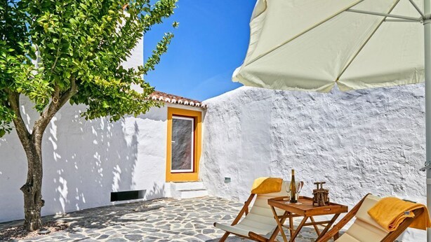 A stone-floored outdoor seating area with two wooden lounge chairs and a table, a tree, and a white wall with a door. - Veiros House / Estremoz II (Veiros)