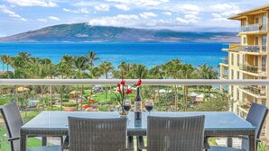 Un balcon avec des fauteuils en osier et une table, donnant sur une plage bordée de palmiers et surmontée, au loin, d’une montagne.