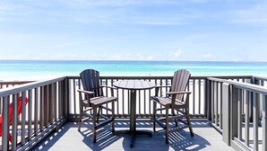 Une terrasse en bois avec deux chaises et une table, donnant sur une plage aux eaux d’un bleu limpide et sur une côte lointaine.