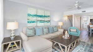 Un salon avec un canapé d’angle beige, deux tables d’appoint avec des lampes, une table basse et un ventilateur de plafond.
