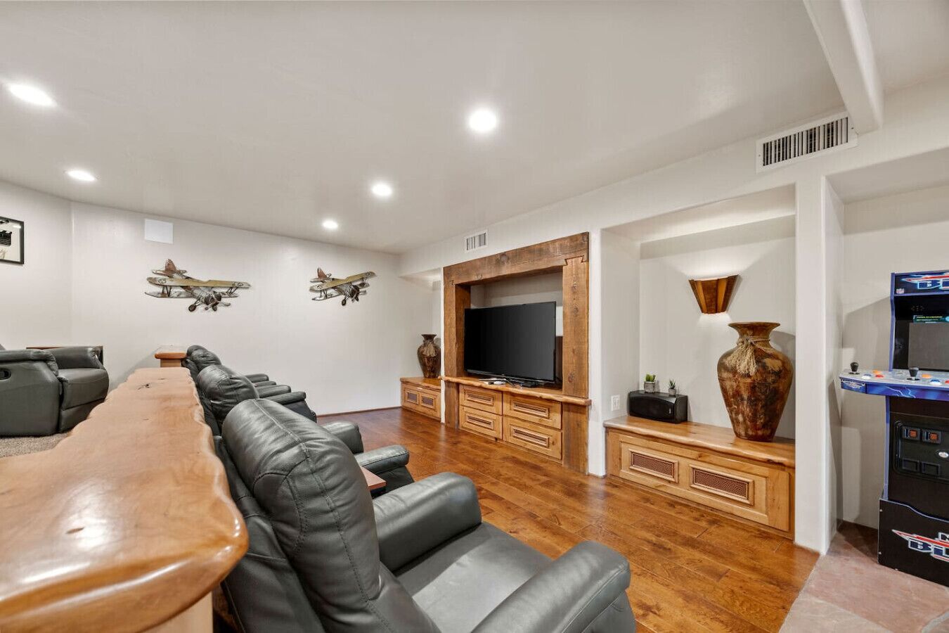 A spacious living room with a long wooden coffee table, leather sofas, a flat-screen TV, wooden cabinets, and decorative items including a large vase and model airplanes.