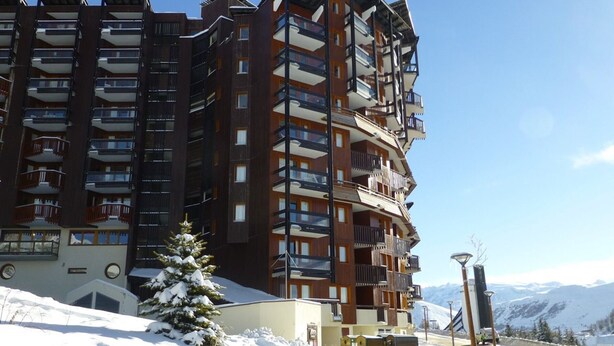 A multi-story hotel with balconies, a snow-covered ground, and a mountainous backdrop. - Cozy 1BR Apartment for 4 with Balcony & Parking, 450m from Lifts – L'Alpe d'Huez (Huez)