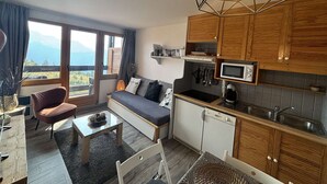 A compact kitchen with wooden cabinets, a microwave, and a stove. There's a sofa and a dining table with a vase of flowers. - Cozy 1BR Apartment for 4 with Balcony & Parking, 450m from Lifts – L'Alpe d'Huez (Huez)