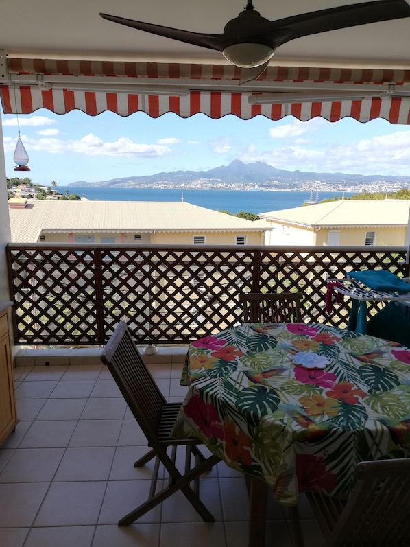 Ein Balkon mit Blick auf das Meer und die Berge, ein Tisch mit einer blumigen Tischdecke und ein Deckenventilator.