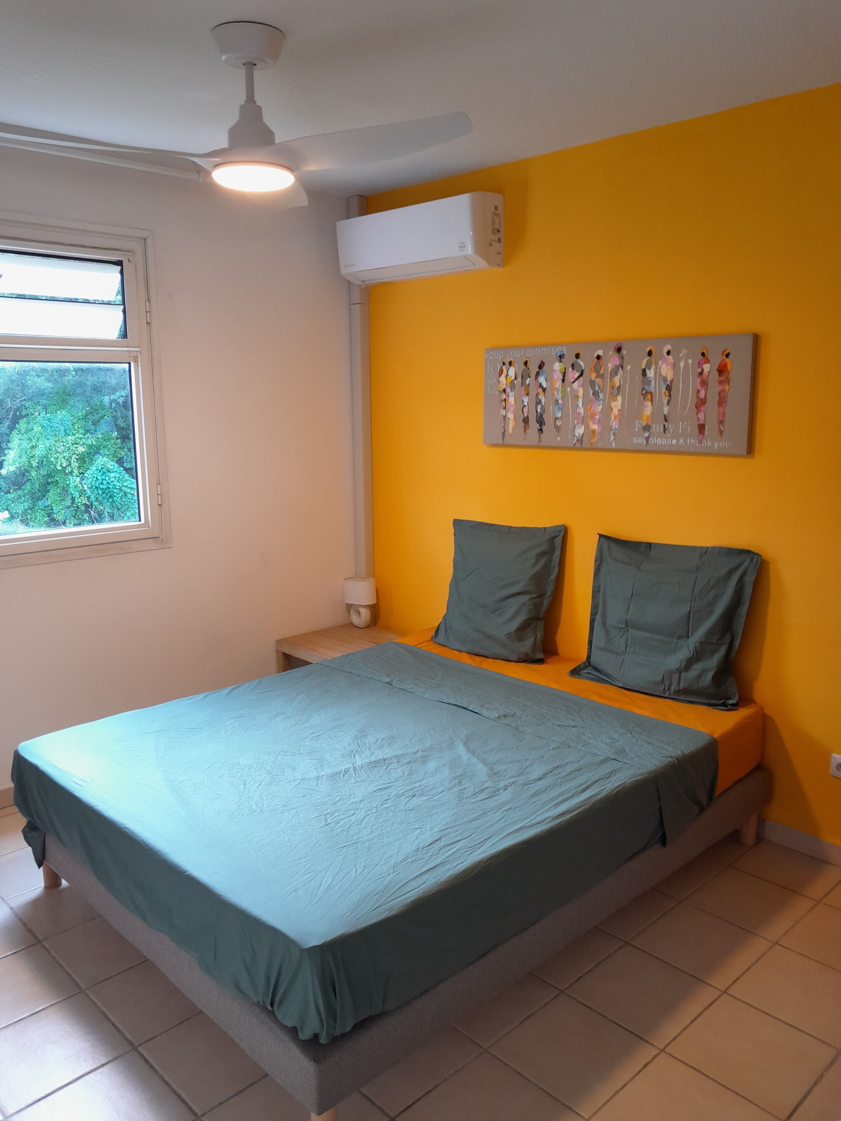 A bedroom with a bed, a ceiling fan, an air conditioning unit, a window, and a wall with a colorful artwork.