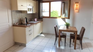 A compact kitchen with a dining area, featuring a window with curtains, a kettle, and a small table with chairs. - Aurore du Lac - Duplex Apartment 4pers. + Studio 2pers. (Sevrier)