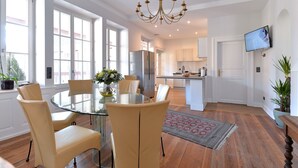 Una cocina moderna con armarios blancos, un refrigerador de acero inoxidable y un horno integrado. Hay un comedor con una mesa de vidrio y sillas color beige.