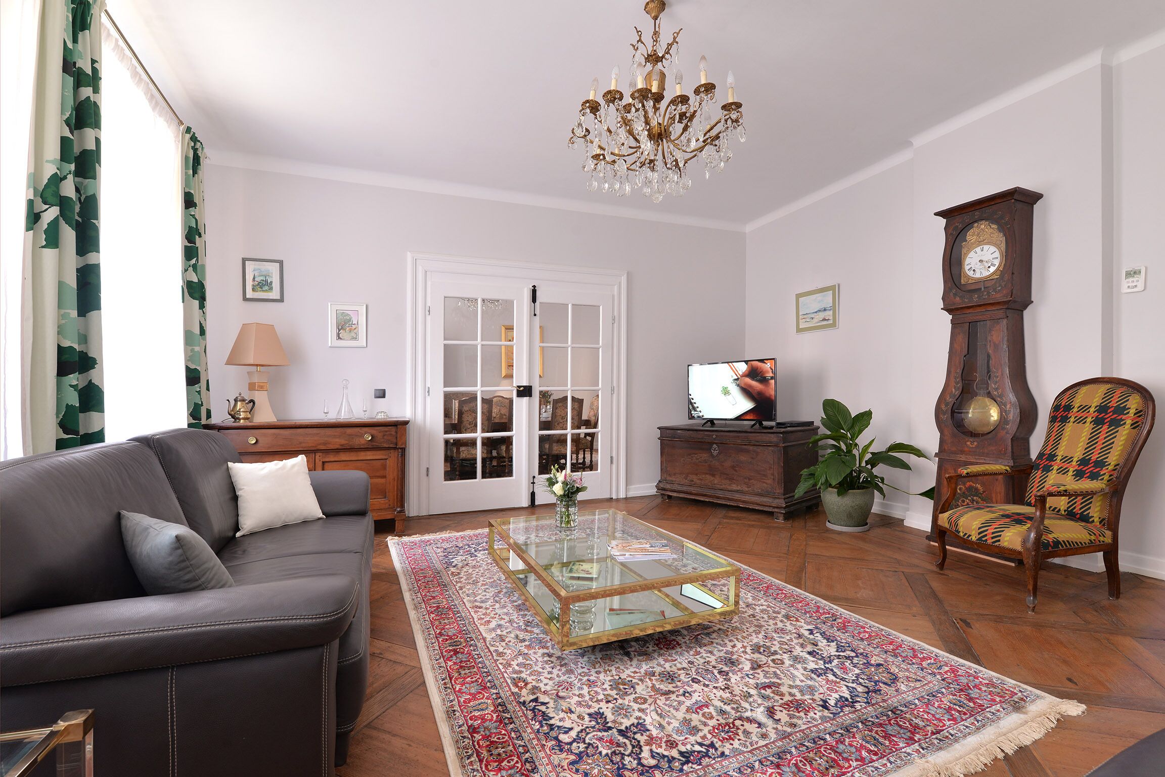 A living room with a leather sofa, a glass coffee table, a patterned rug, a wooden floor, a chandelier, a clock, a television, and a potted plant.
