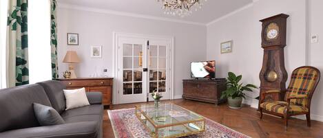 A living room with a leather sofa, a glass coffee table, a patterned rug, a wooden floor, a chandelier, a clock, a television, and a potted plant.