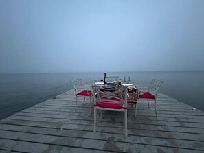 Een houten steiger die uitsteekt in een rustig waterlichaam, met een tafel voor twee personen en twee stoelen met rode kussens.