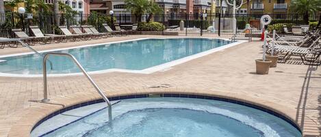 Una piscina con sillas de descanso y un jacuzzi, rodeada de una valla y edificios.