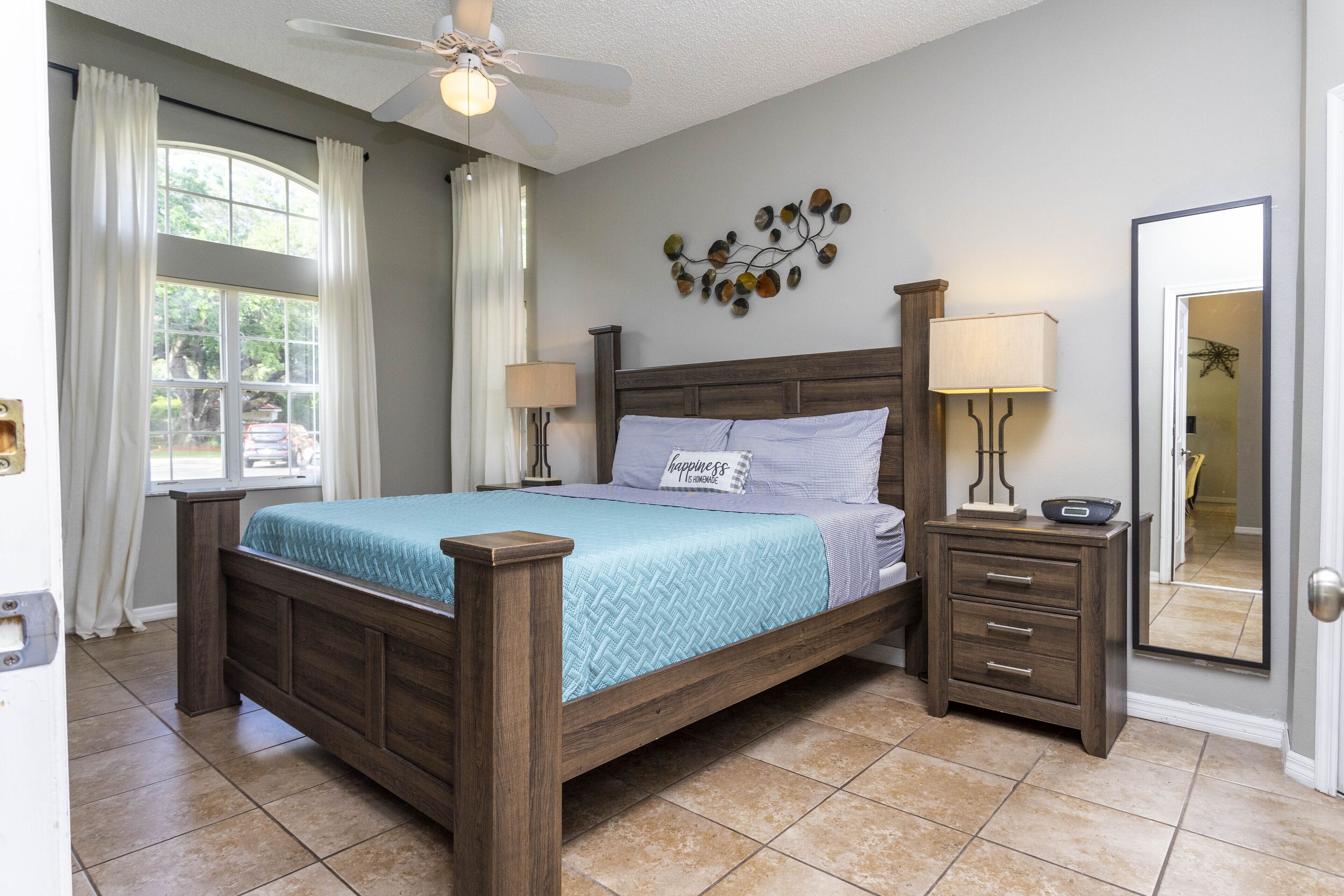 A bedroom with a wooden bed, two bedside tables, a nightstand, a ceiling fan, a window with curtains, and a wall-mounted decoration.