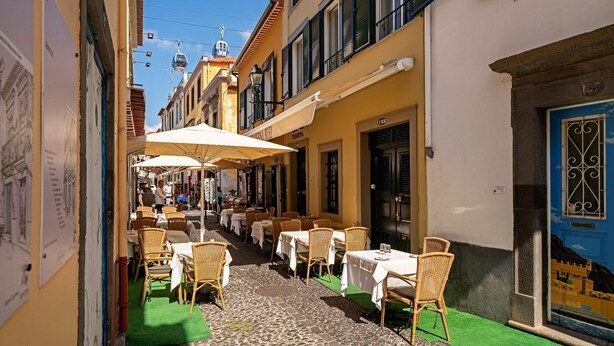 A narrow street with outdoor seating, wicker chairs, and tables under umbrellas.