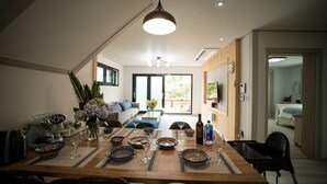 Un comedor con una mesa de madera dispuesta para cuatro personas, rodeada de sillas. Se observa una mesa con mantel individual, cuencos, copas de vino y una botella. La habitación presenta un diseño moderno, con una lámpara de techo, un sofá y una ventana con persianas.