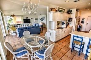 Uma cozinha moderna com armários brancos, um espaço de jantar com cadeiras de vime e uma sala de estar com um sofá azul.
