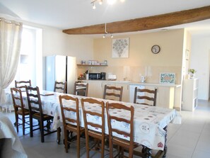 A dining area with a table set for six, wooden chairs, a kitchen in the background with appliances, and a clock on the wall.