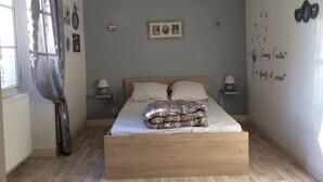 Une chambre à coucher avec un lit en bois, deux tables de chevet, une fenêtre avec des rideaux et des éléments décoratifs sur les murs, notamment un tableau encadré et une œuvre d’art murale avec du texte.