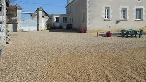 Un bâtiment avec une allée en gravier, un petit jardin agrémenté de plantes en pot et une table avec des chaises à l’extérieur.