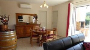 Uma sala de estar com sofá de couro preto, mesa de jantar de madeira com cadeiras, área de cozinha com armário de madeira e uma janela grande com vista para o verde lá fora.