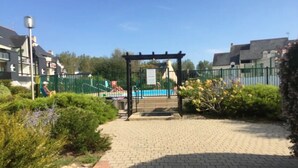 Uma trilha pavimentada leva a uma pérgola de madeira com uma placa, cercada por vegetação e uma área cercada com uma piscina ao fundo.