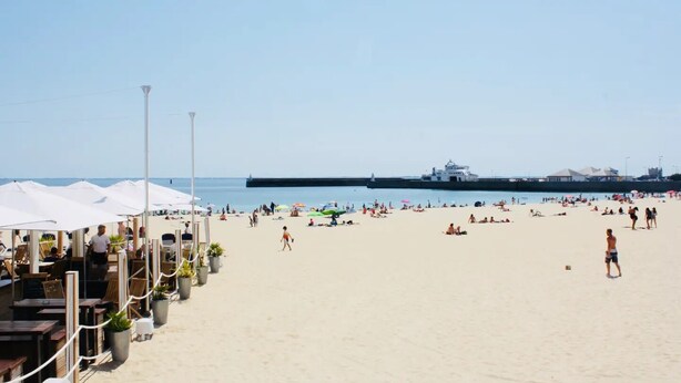 Uma praia com guarda-sóis, espreguiçadeiras e um cais ao fundo.