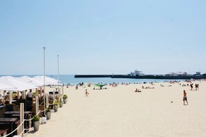 En strand med parasoller, solstolar och en brygga i bakgrunden.