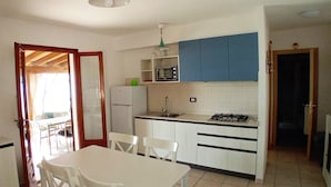 Une cuisine compacte avec des meubles blancs, un four à micro-ondes, un évier et une cuisinière à gaz. On y trouve une table à manger avec des chaises et un téléviseur fixé au mur.