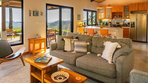 A modern living room with a grey sofa, wooden coffee table, and a view of a kitchen and mountains through large windows. - Stunning house - your oasis in Samara, 3 minutes to beach (Sámara)